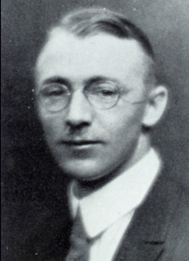 Image description: black and white headshot of Joseph Gilchrist wearing a suit with glasses.