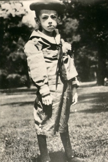 black and white photograph of young Teddy Ryder standing outside in shorts, a top with a tie, and a hat. He is very thin.