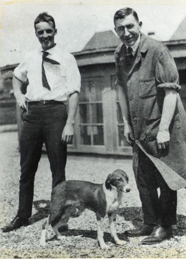 Best standing on the left wearing a short sleeve white shirt with tie blowing in the wind. He is smiling at the camera and has a hand on his hip. Banting is standing on the right in a lab coat and slightly hunched over. There is a black and white dog standing between them.