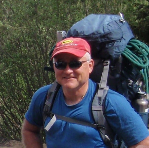Michael smiling and wearing sunglasses, a cap, and a backpack while hiking.