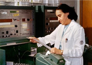 Yalow is standing in front of large machines and wearing a white coat. She is looking at a long strip of paper.