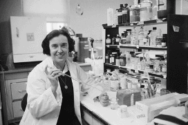 Yalow sitting at a lab bench holding a piece of lab equipment and smiling at the camera. She is wearing a white lab coat and has bottles stacked on a shelf behind her.